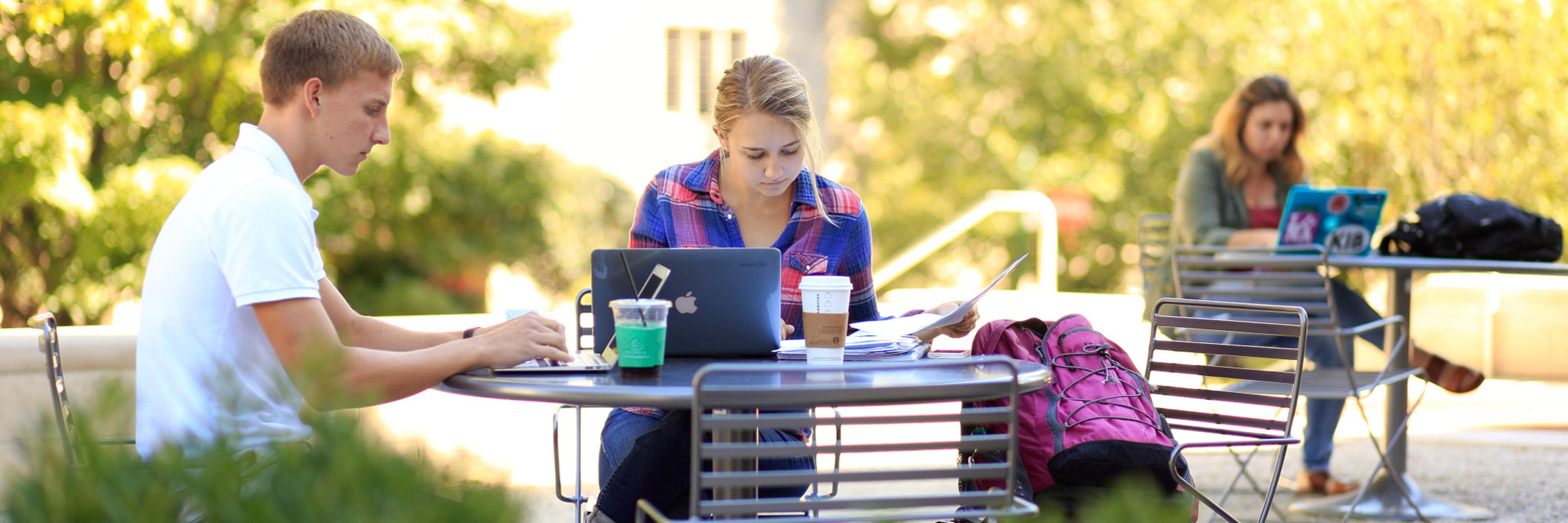 Three students sit outdoors working on their laptops